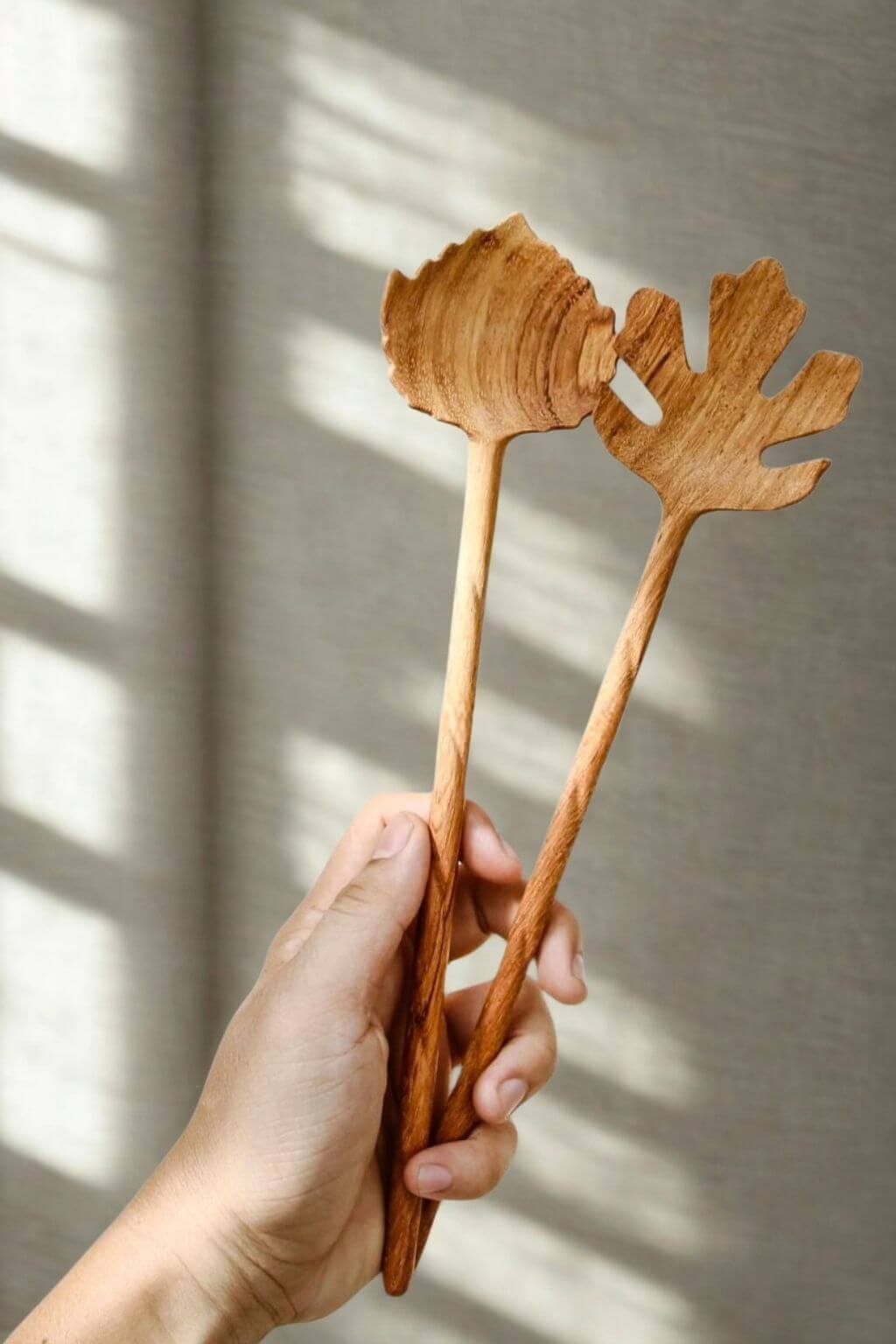 A hand holding two leaf-shaped wooden serving utensils with long handles, shown against a neutral textured wall with soft window shadow light.