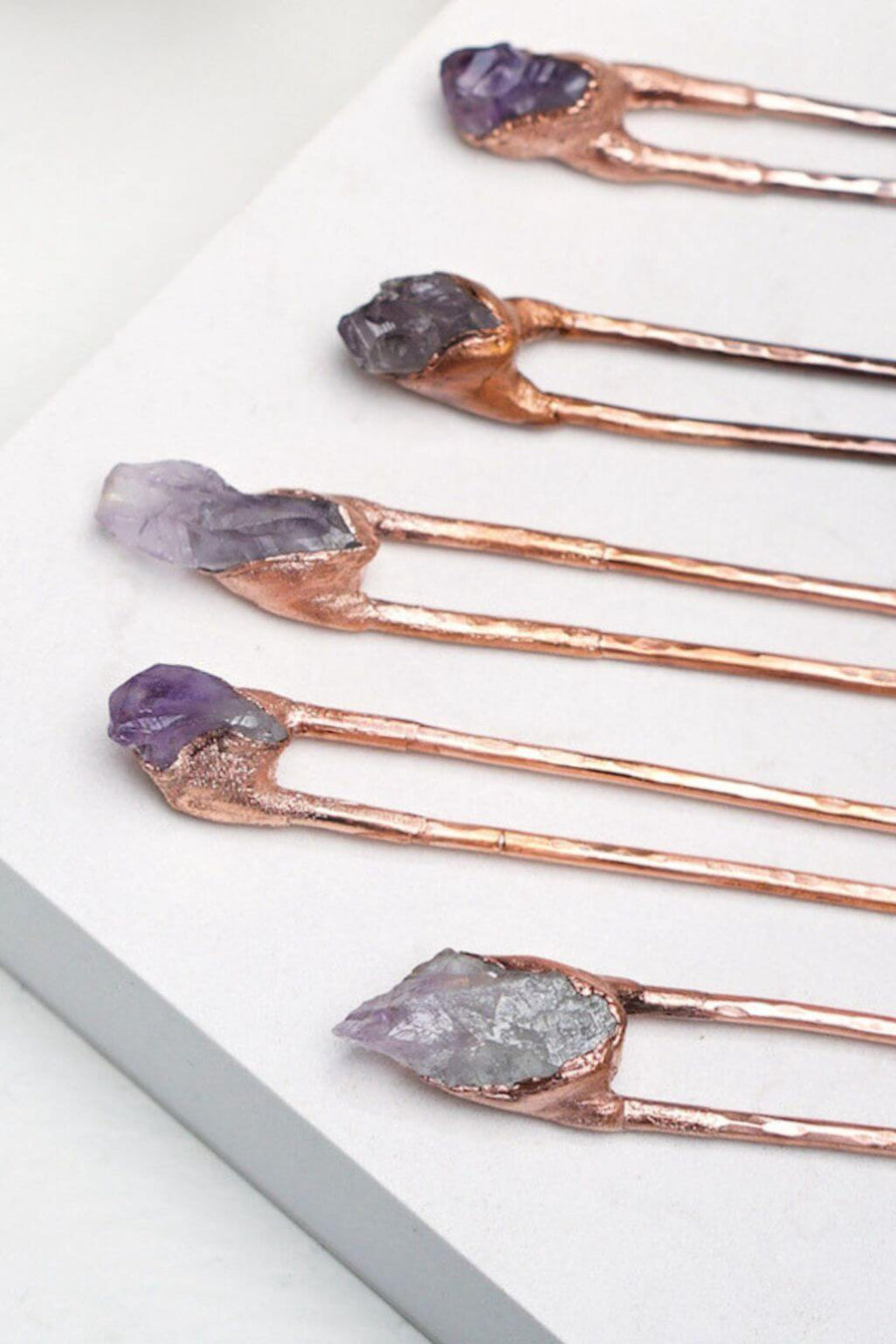 Five copper hair pins with natural crystal tops in purple, gray, and lavender tones, arranged diagonally on a white surface.