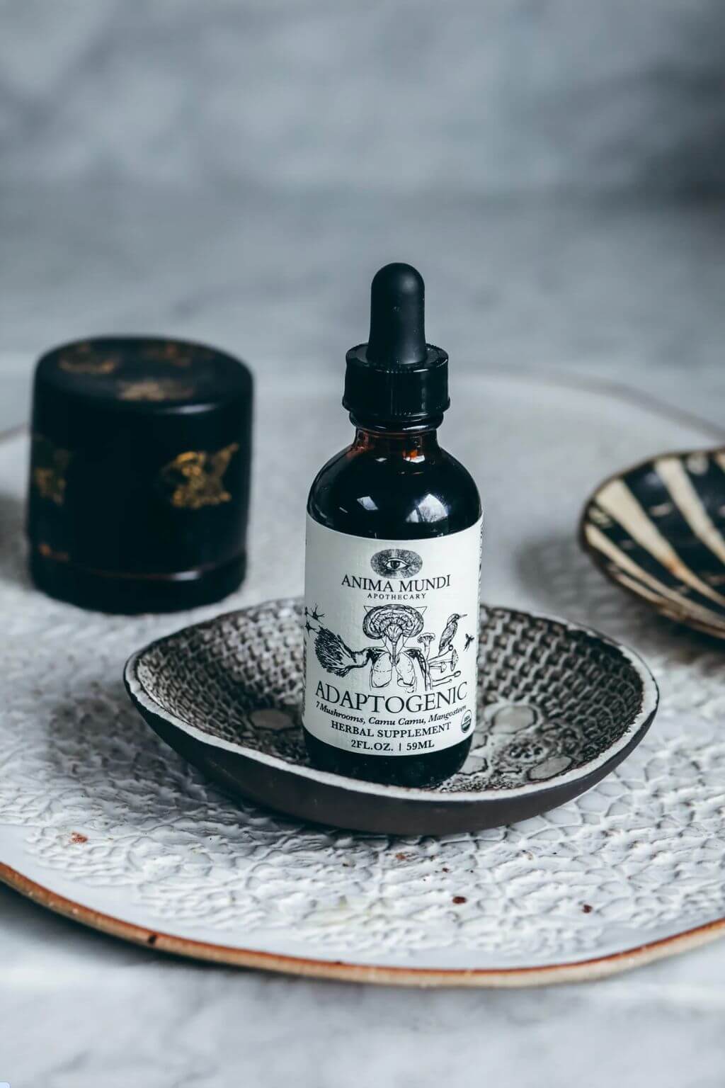 Anma Munde Adaptogenic herbal supplement in an amber glass dropper bottle, styled on a textured ceramic dish with a black container in the background.