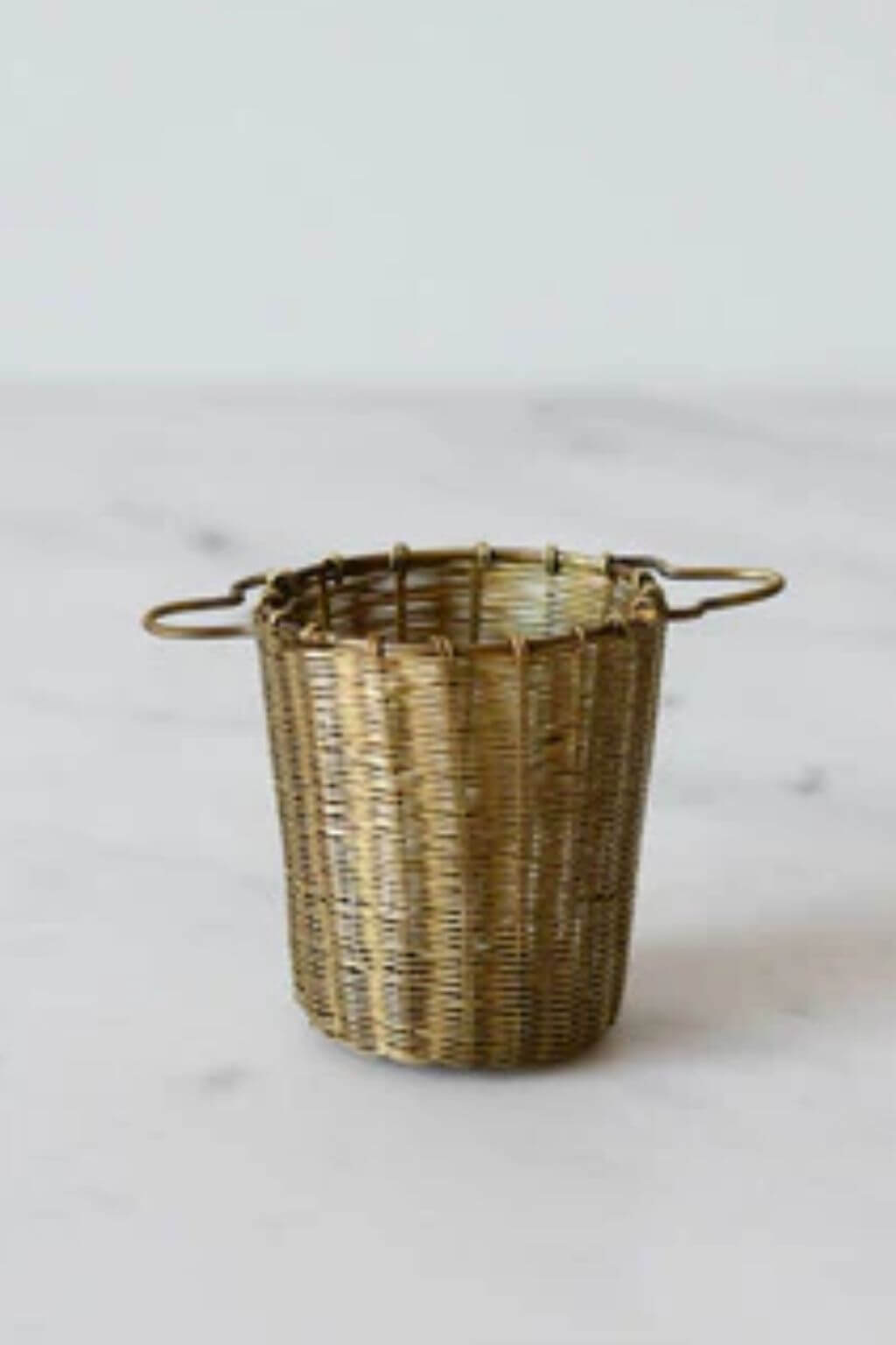 A woven tea basket with two loop handles, made from brass, set on a light marble surface against a white background.