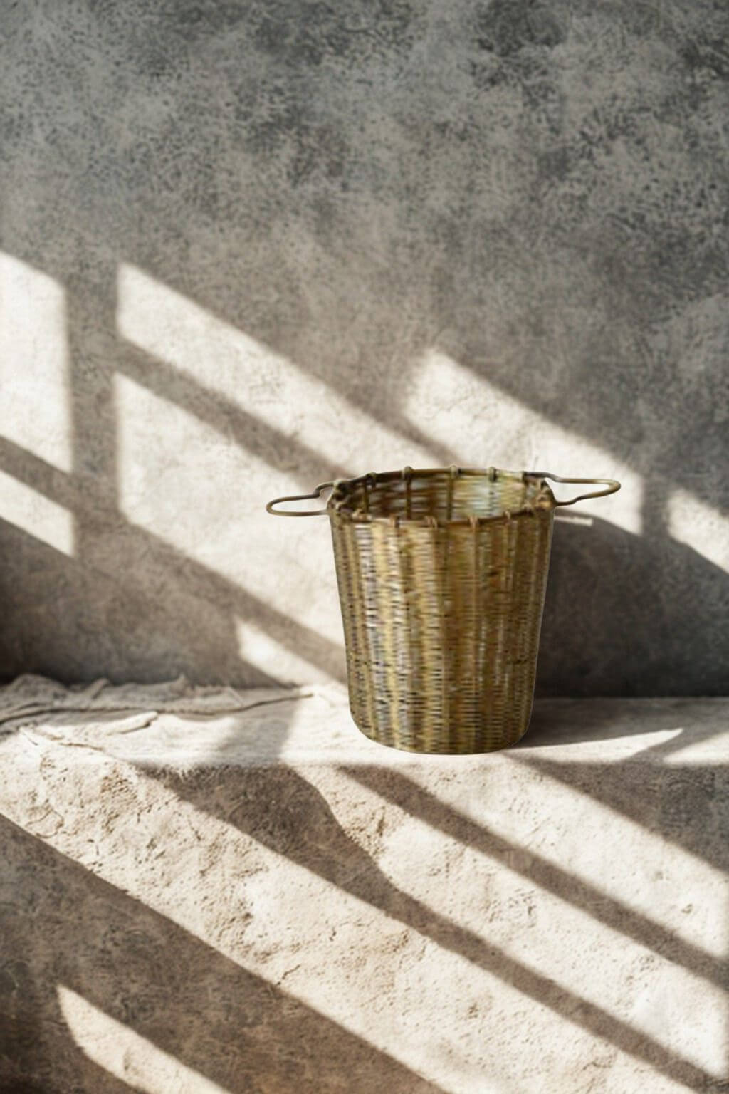 A small woven brass infuser basket with two loop handles sits on a rough stone surface against a textured concrete wall, lit by strong diagonal shadows.