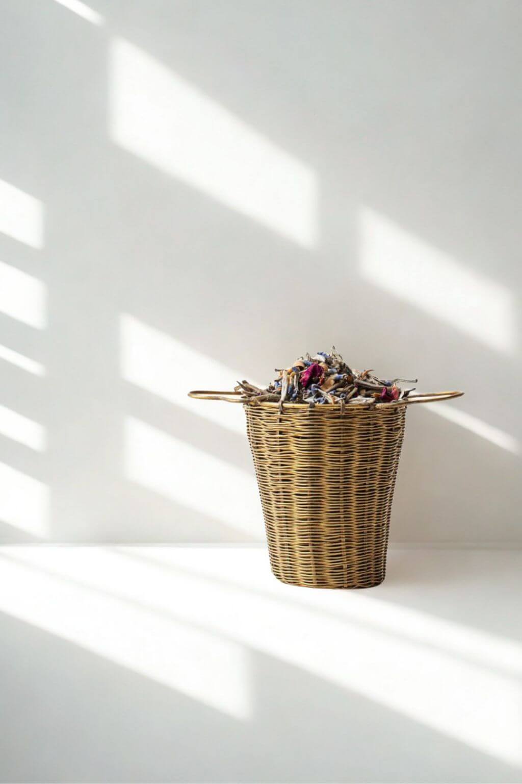 A natural woven brass infuser basket with two side handles sits on a white surface against a white wall, lit by soft diagonal window shadows.