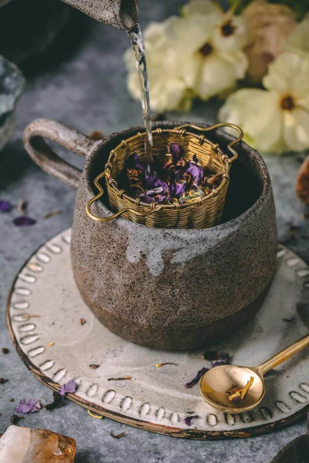 Tea being poured into a brass tea infuser inside a ceramic cup with dried flowers