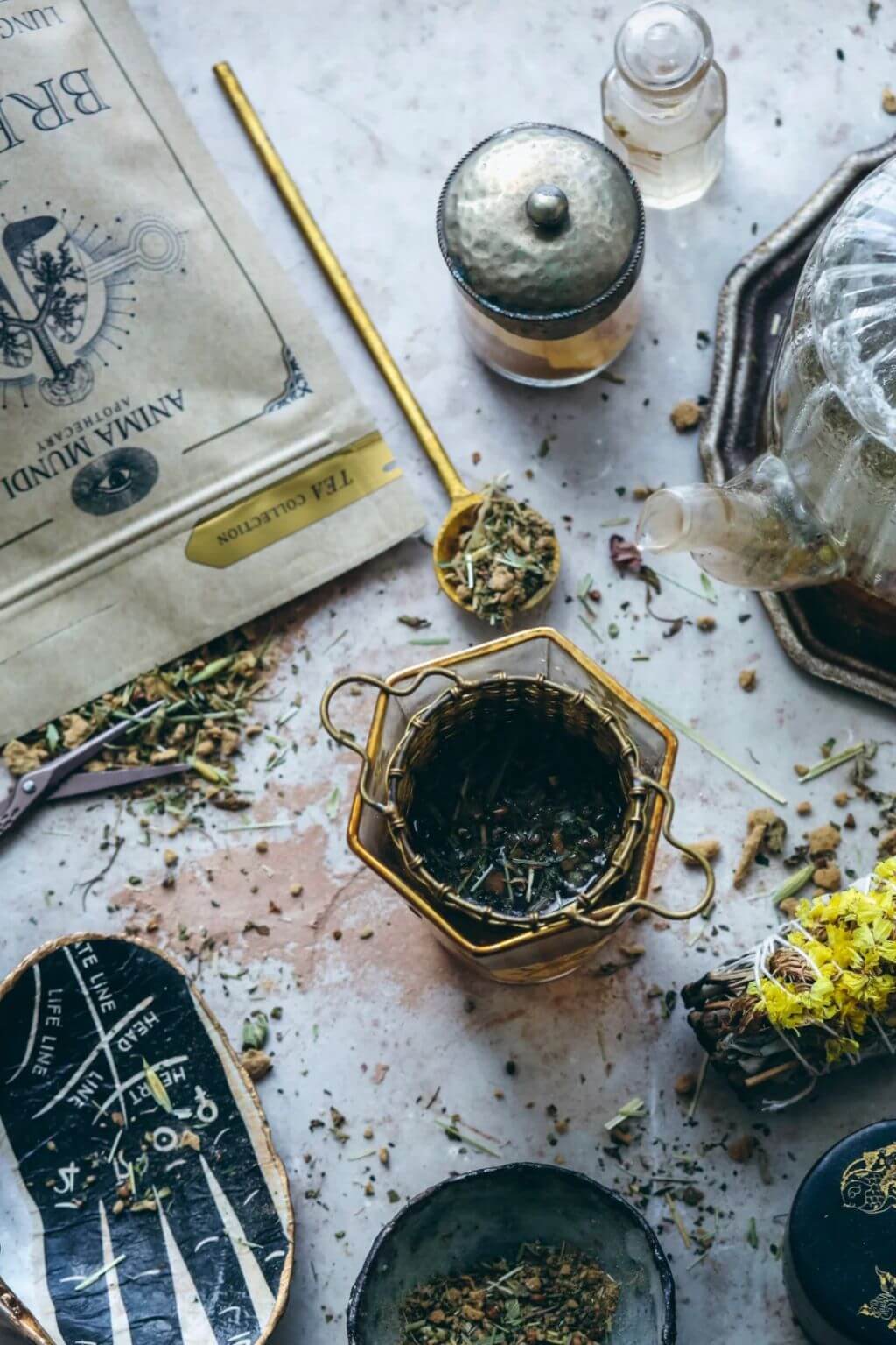 A styled flat-lay of tea accessories and loose leaf tea on a light stone surface, including a branded tea pouch, a hexagonal metal tea infuser, a glass teapot, a small glass jar, a gold spoon, and bowls with loose tea.