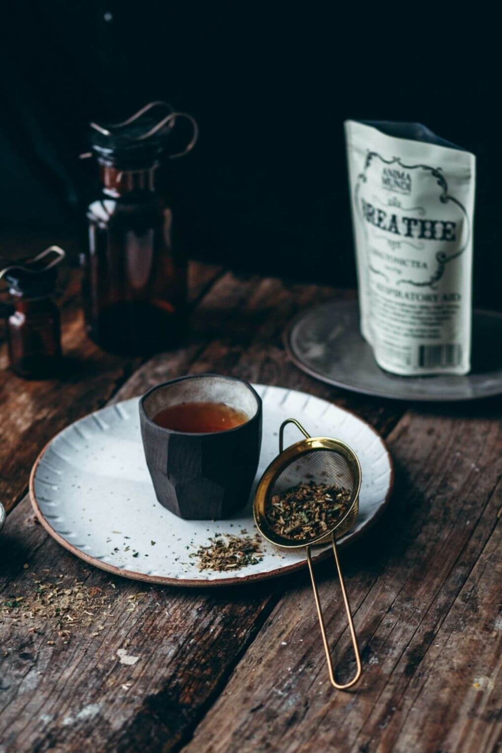 A dark ceramic cup filled with tea sits on a speckled white plate next to a brass mesh strainer holding loose leaf tea, with a white pouch labeled 'BREATHE' standing in the background on a rustic wooden table.