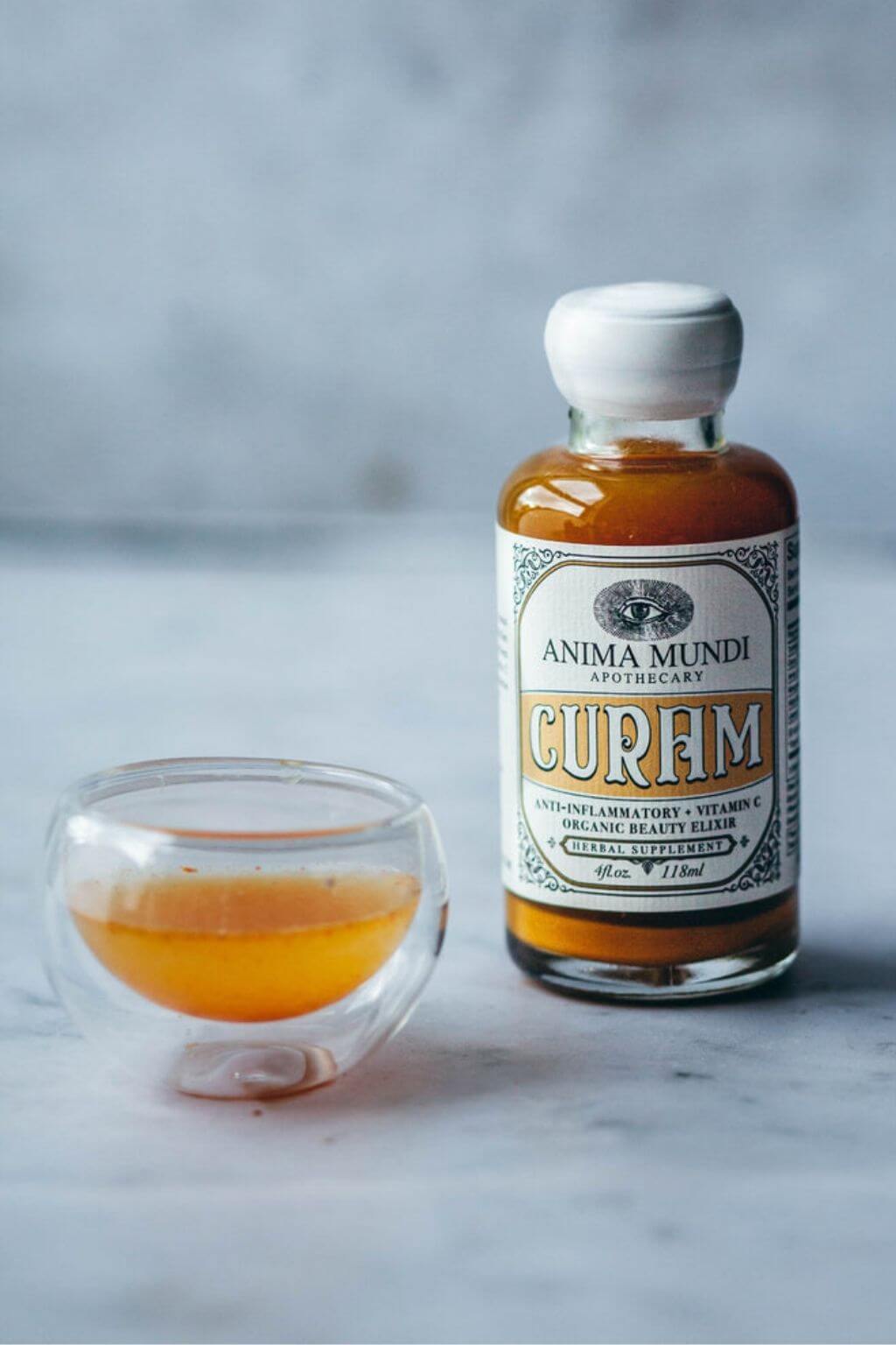 Bottle of Anima Mundi Curam with a glass containing the orange tinted elixir on a marble surface.