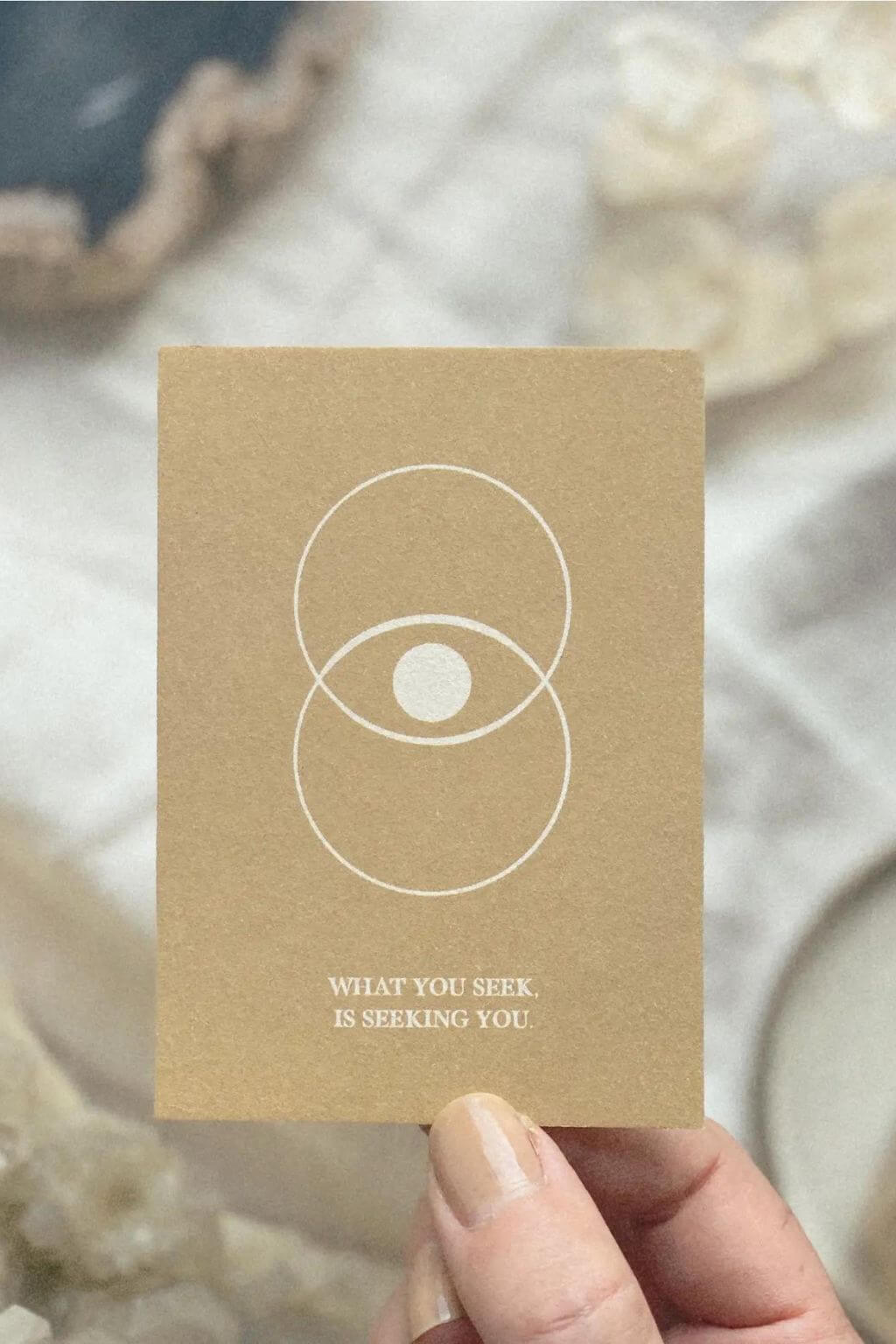 Hand holding an affirmation card reading What You Seek Is Seeking You against a soft neutral background