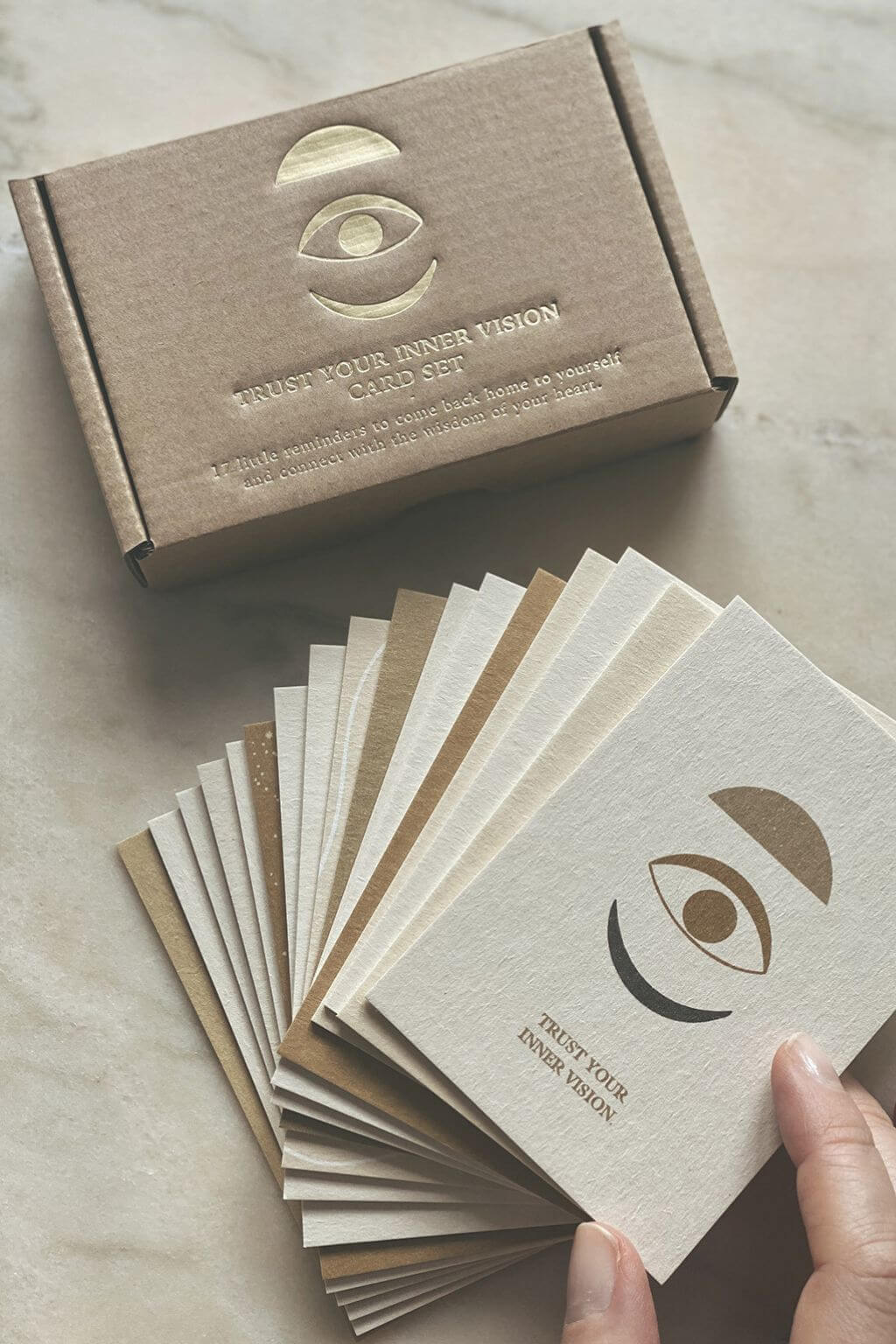 A kraft paper box labeled 'Trust Your Inner Vision Card Set' sits on a light marble surface with a hand holding a fanned-out selection of matching cream and tan cards. The cards and box feature an embossed eye logo and the text 'TRUST YOUR INNER VISION'.