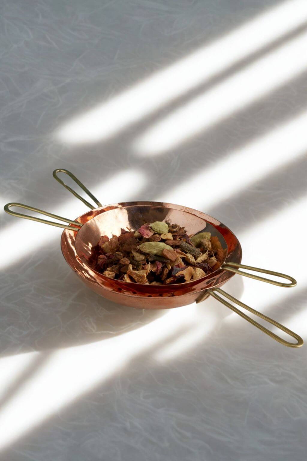 Copper tea strainer with brass handles holding dried herbs and tea leaves on a rice paper background with gentle light and intricate shadows