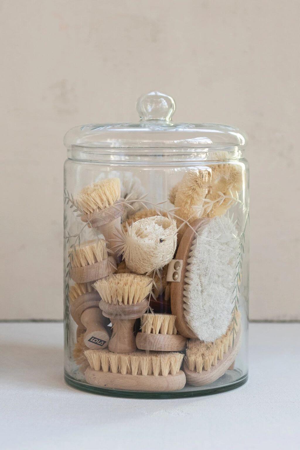 A clear glass jar with a lid containing an assortment of natural fiber and wood bath brushes, set against a neutral beige background.