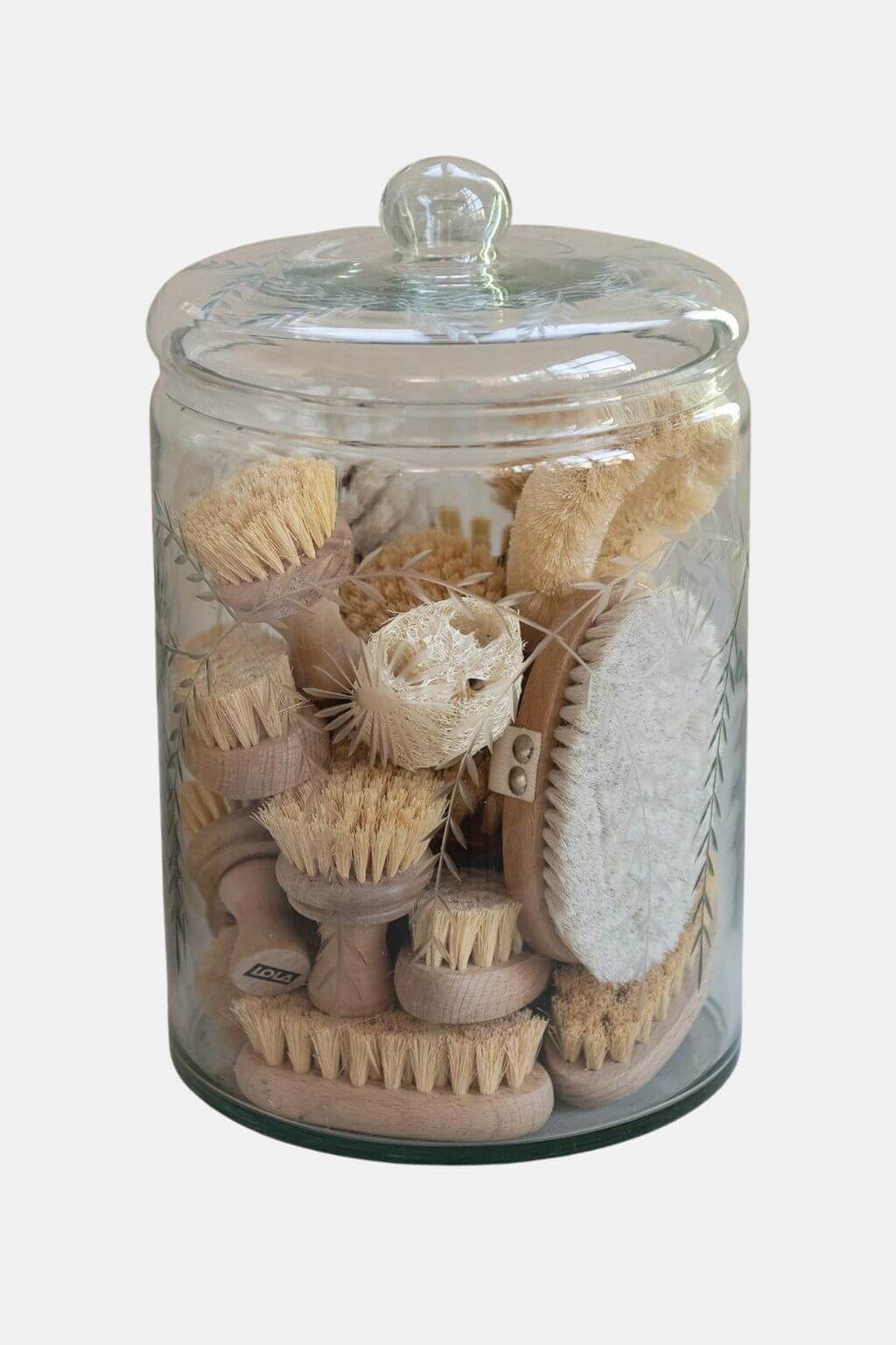 A clear glass jar with a matching glass lid, filled with assorted natural fiber and wood body brushes, shown against a light gray background.
