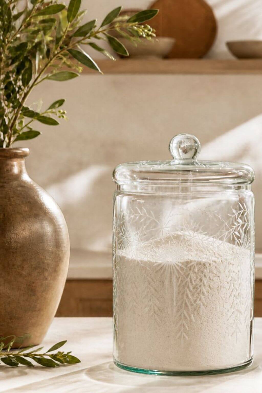 A clear glass storage jar with a lid, filled with flour, placed on a countertop beside a ceramic vase with greenery in a softly lit kitchen