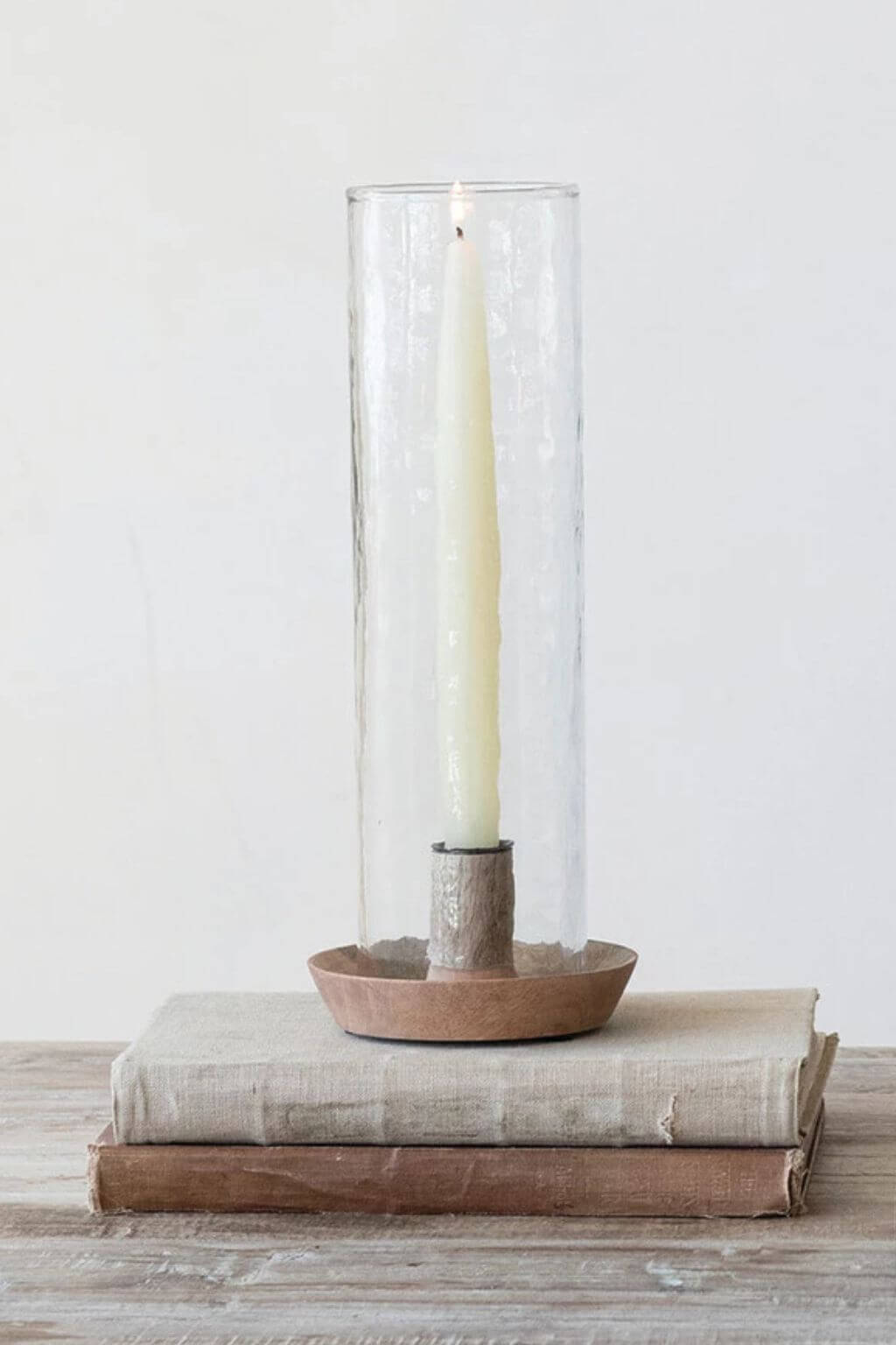 A tall clear glass candle holder with a white taper candle, set on a small round wooden base and resting on stacked books against a plain light background.