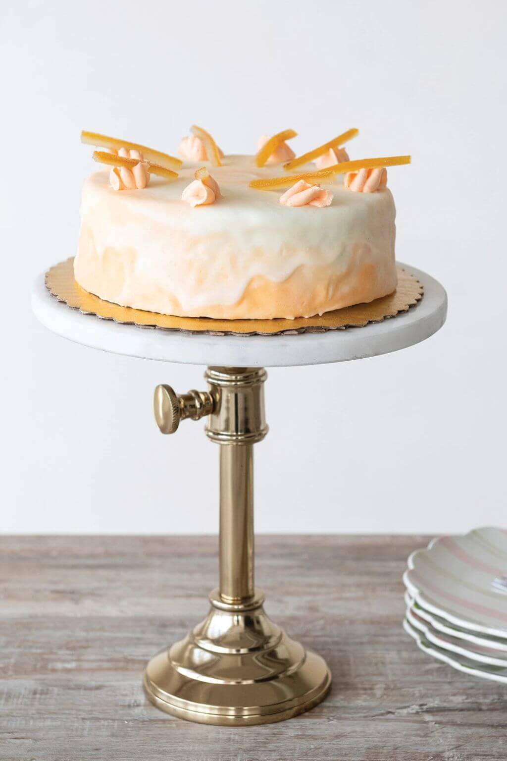 A round frosted cake with peach-colored frosting and orange garnish sits on a white marble-top pedestal stand with a gold base, set on a rustic wooden table.