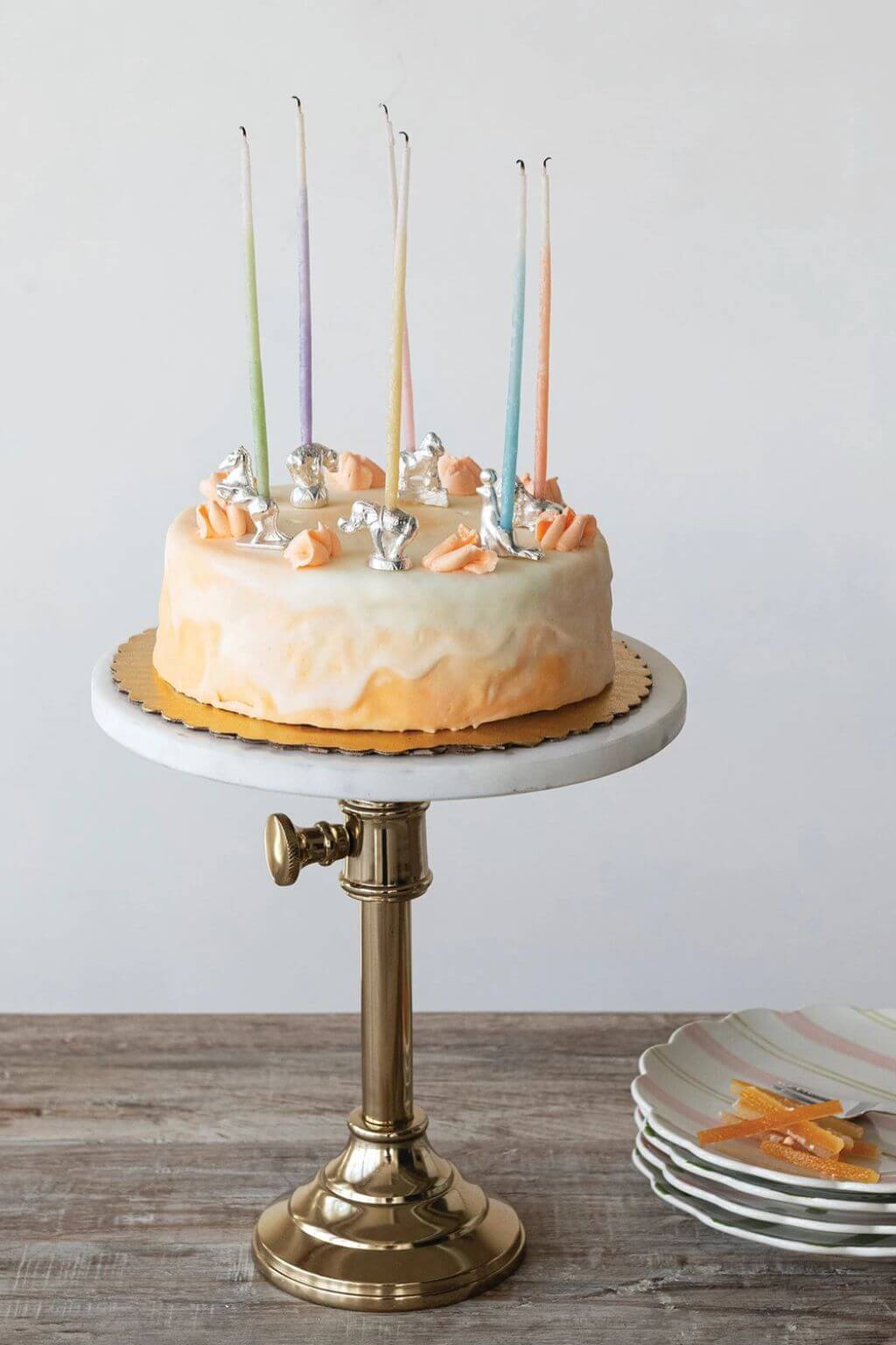A frosted birthday cake with orange-peach icing, decorative silver foil pieces, and six tall multicolored taper candles on a white marble and brass cake stand, set on a wooden table with plates in the background.