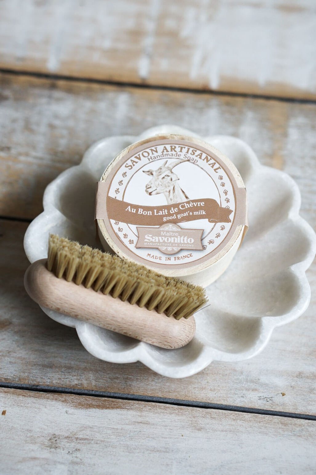 A small round tin of artisanal goat milk soap with a goat logo and French text, placed on a white scalloped dish with a wooden-handled brush on a rustic whitewashed wood surface.