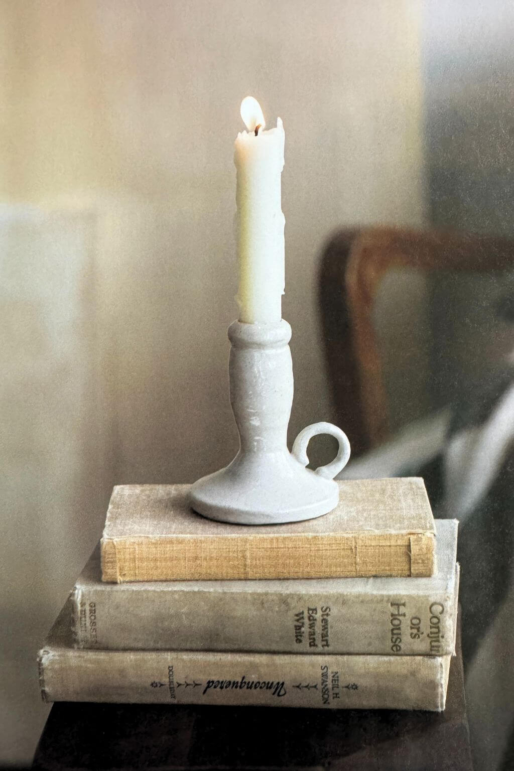 A white ceramic candlestick holder with a loop handle, holding a lit white candle, resting on a stack of vintage books.