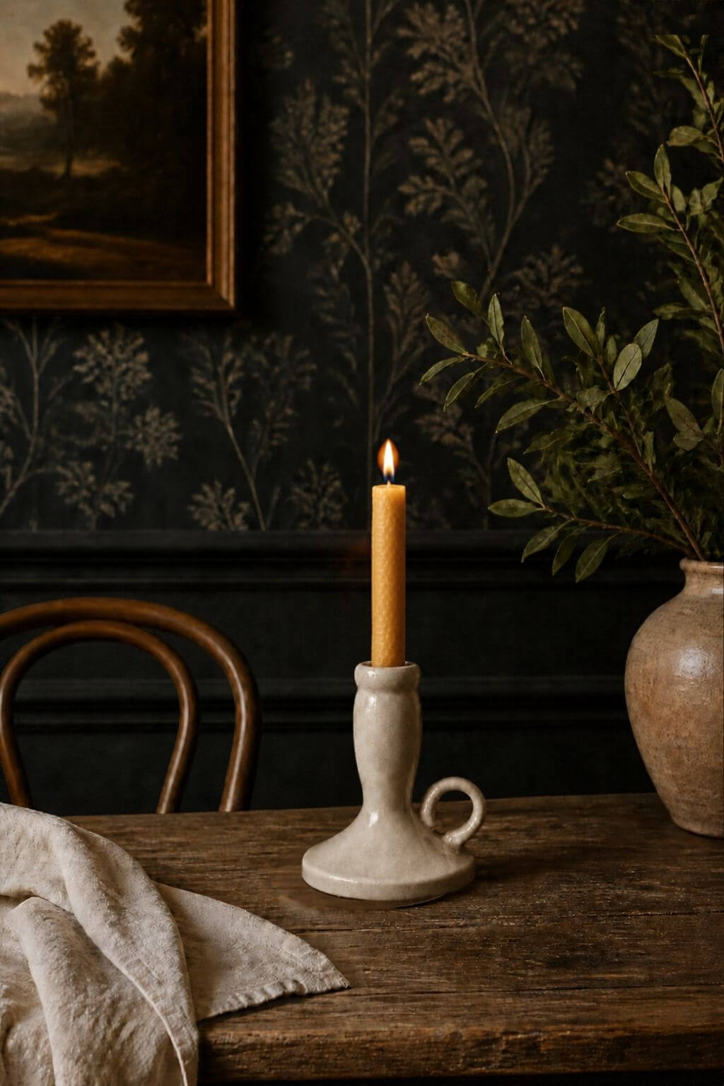A lit taper candle in a matte ceramic candlestick holder with a loop handle, set on a rustic wooden table in a dark, vintage-style room with floral wallpaper, a chair, and a vase.