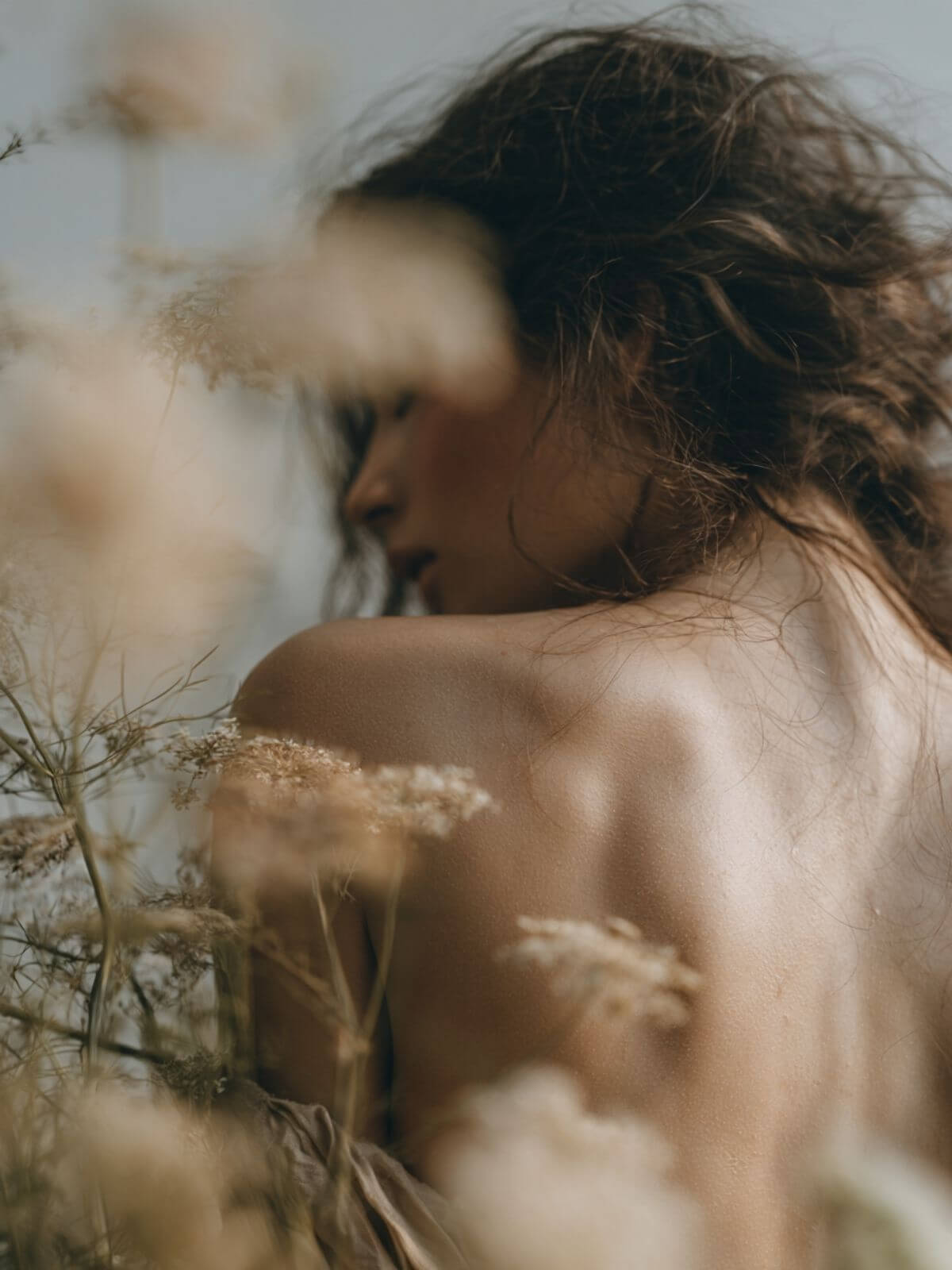 A close-up lifestyle photograph of a woman outdoors, seen from behind with her dark hair blowing in the wind, surrounded by soft-focus beige wildflowers.