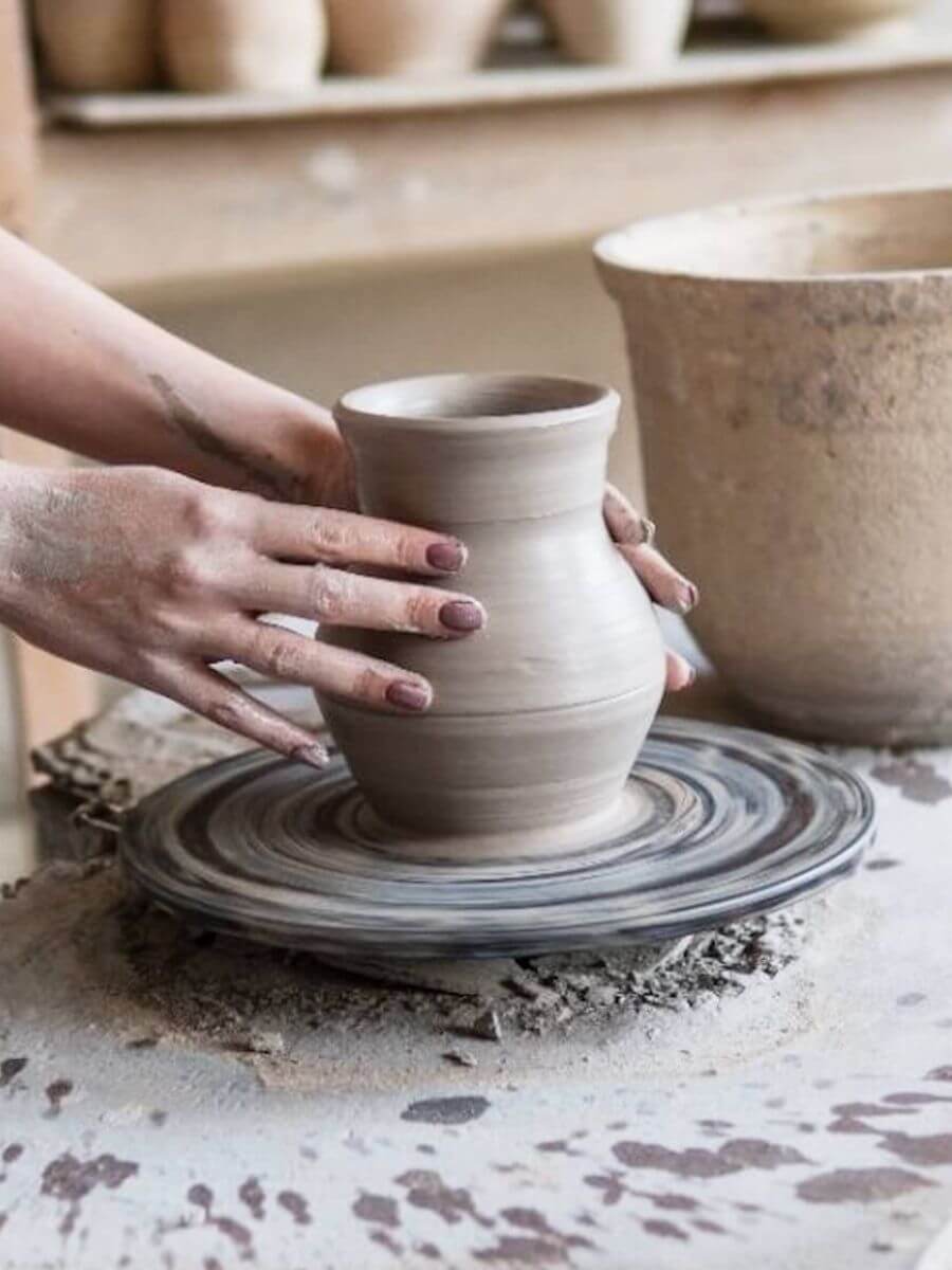 A person's hands shaping a small handmade clay pot on a pottery wheel, with a larger clay vessel in the background.