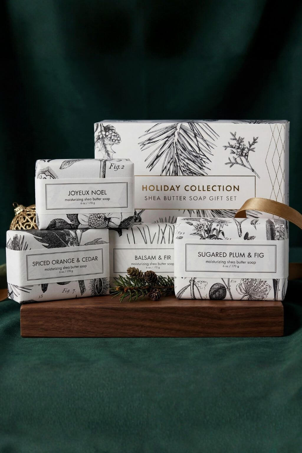 Fig 2 Holiday Collection shea butter soap gift set arranged on a wooden tray against a dark green fabric backdrop. The set includes five individually wrapped soaps with botanical illustrations on the packaging: Joyeux Noel, Spiced Orange & Cedar, Balsam & Fir, Sugared Plum & Fig, and the main Holiday Collection box.