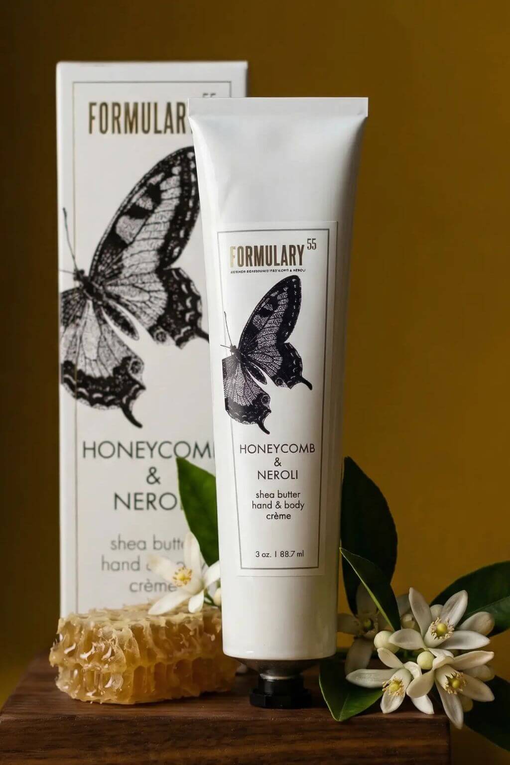 A white tube of Formulary Honeycomb & Neroli hand and body creme stands in front of its matching product box, styled on a wooden surface with a honeycomb piece, green leaves, and white flowers against a warm mustard-yellow background.