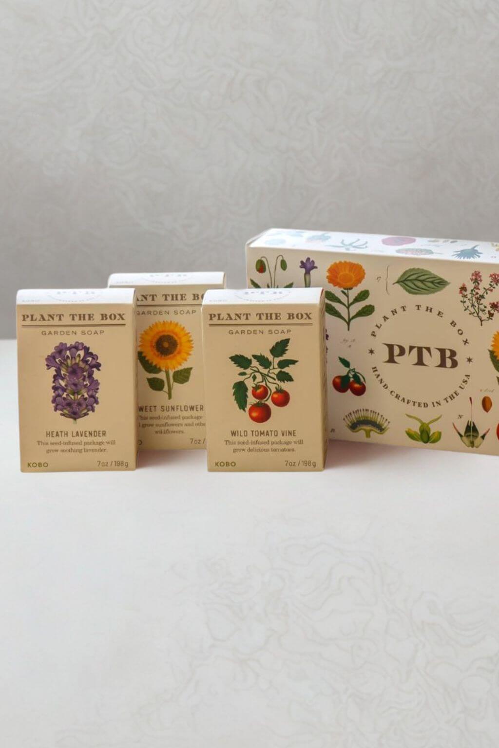 Three small botanical-themed soap boxes in front of a larger matching box on a light patterned surface. The front labels read Plant the Box, with illustrated flowers and fruits and scent names including Lavender, Wild Sunflower, and Wild Tomato Vine.