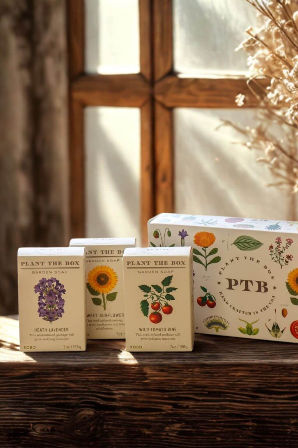 Three small Plant The Box bar soaps arranged on a rustic wooden surface in front of a sunlit window. The boxes are cream-colored with botanical illustrations and labels for lavender, mint sunflower, and wild onion.
