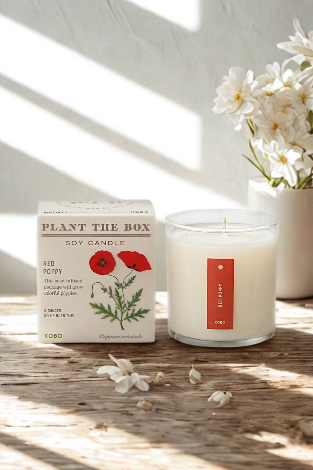A soy candle in a clear glass jar sits beside its white box on a rustic wooden surface. The box reads "PLANT THE BOX" and "RED POPPY," with a red poppy illustration.
