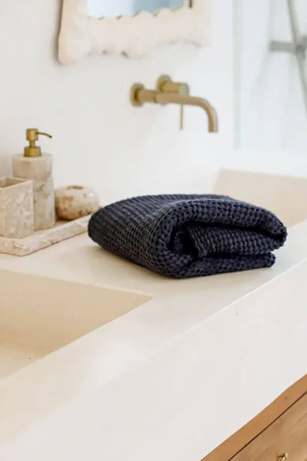 A folded dark navy blue waffle-knit washcloth resting on a beige stone bathroom counter next to a brass faucet and soap dispenser.