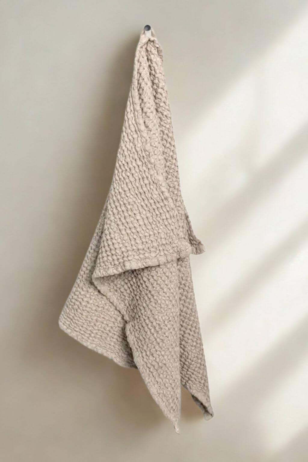 A textured, chunky knit sand towel hanging from a small wall hook against a warm off-white wall with soft diagonal shadows.