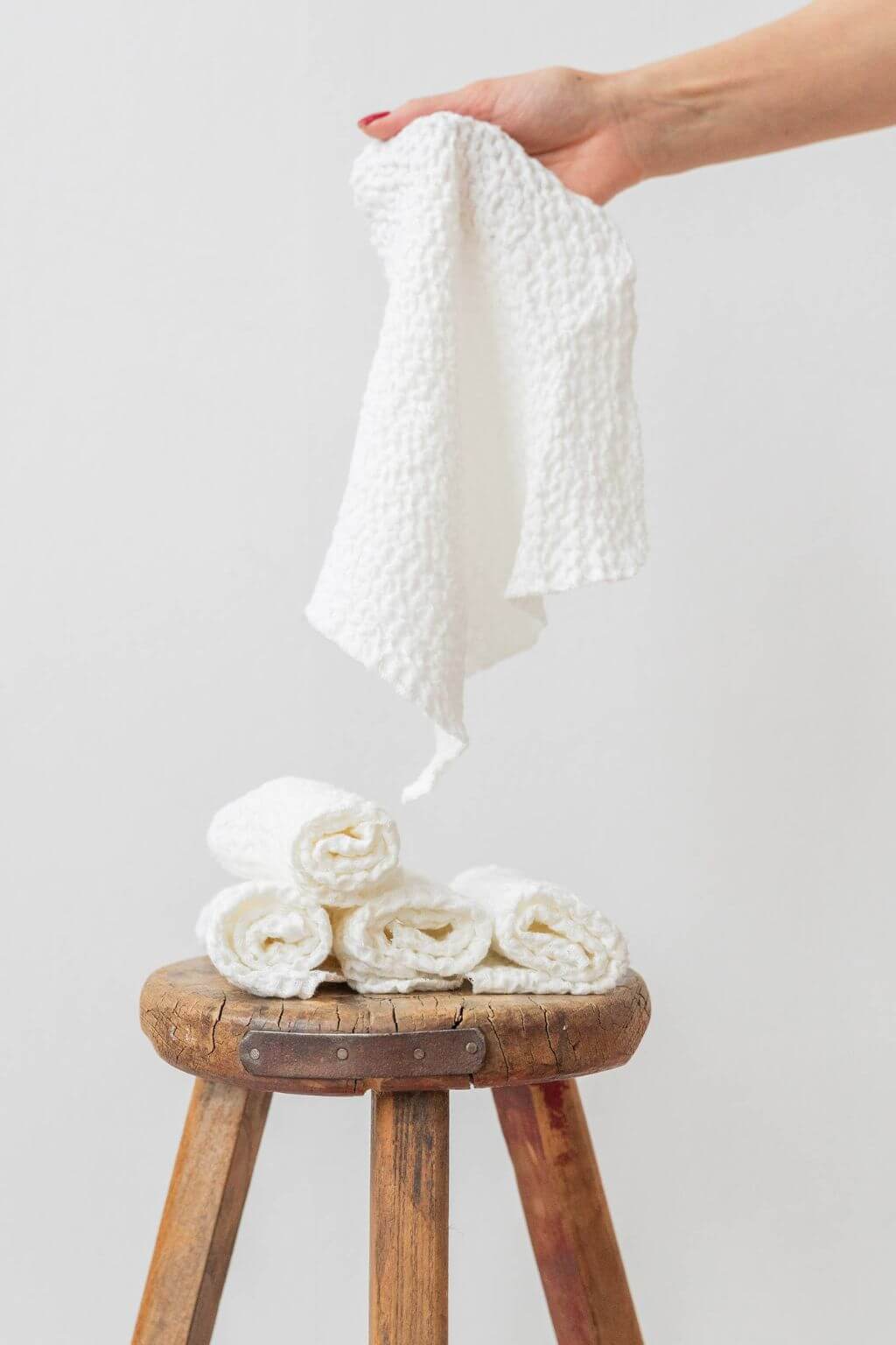White waffle textured fabric being held above a wooden stool with folded white washcloths resting on it.