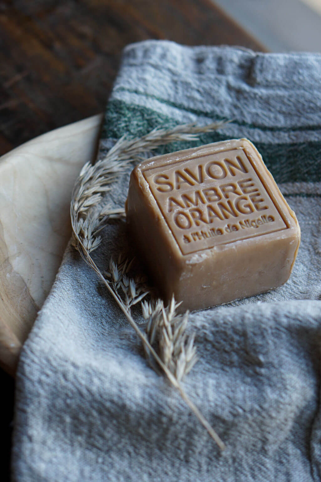 A rectangular bar of Savon Ambre Orange soap resting on a textured gray towel, styled with dried wheat stalks and a wooden bowl in the background.