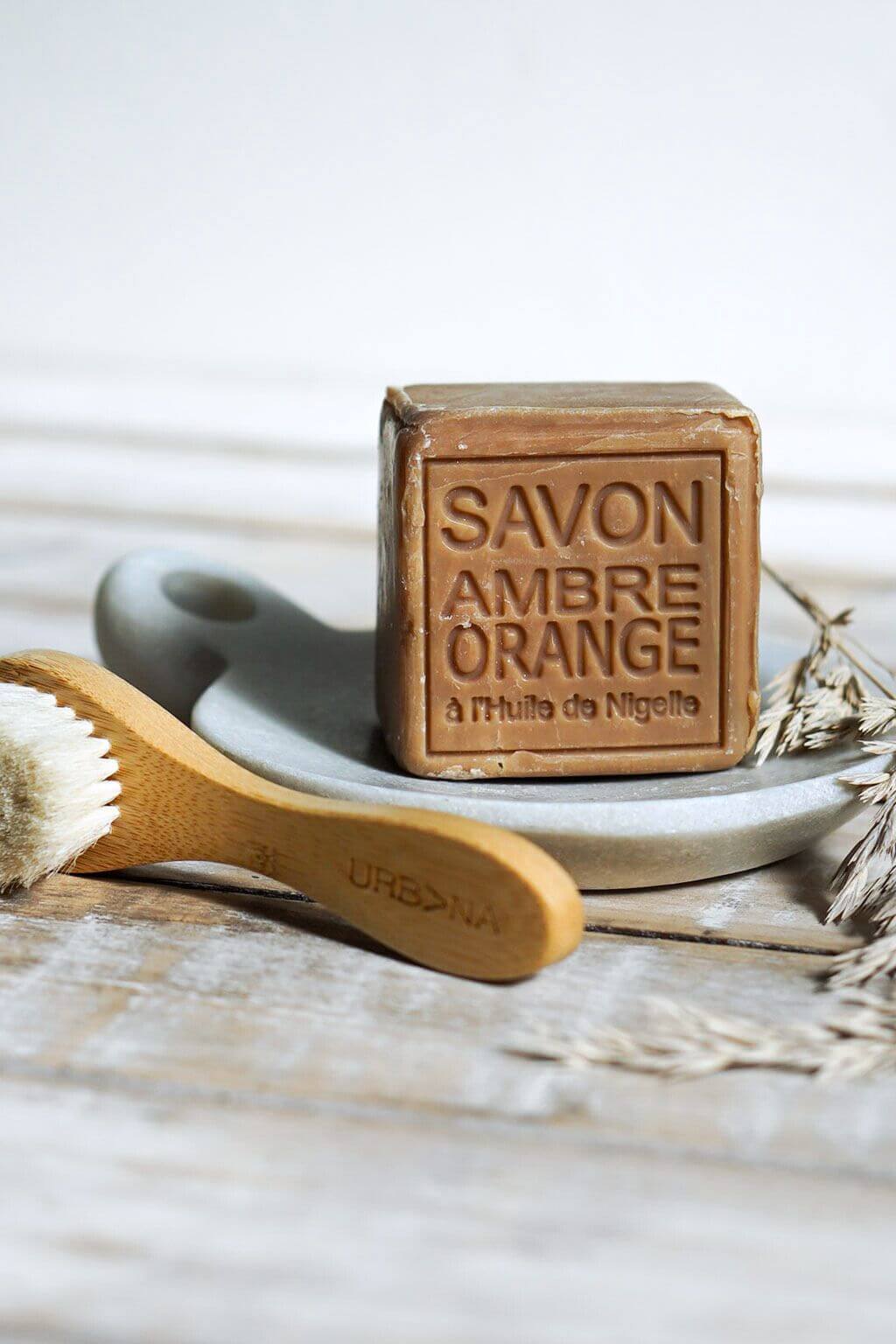 Bar of Savon Ambre Orange soap on a stone plate with a wooden brush, on a light wooden surface.
