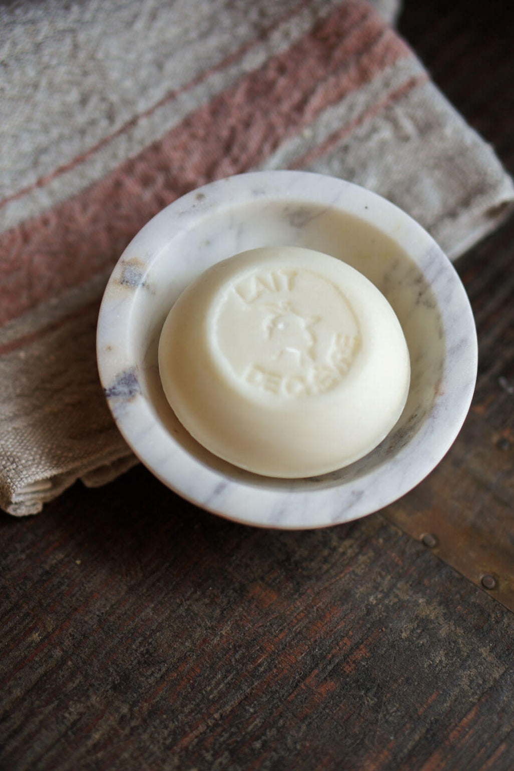 A round bar of off-white soap resting in a small white marble soap dish, placed on a rustic dark wooden surface with a striped cloth underneath.