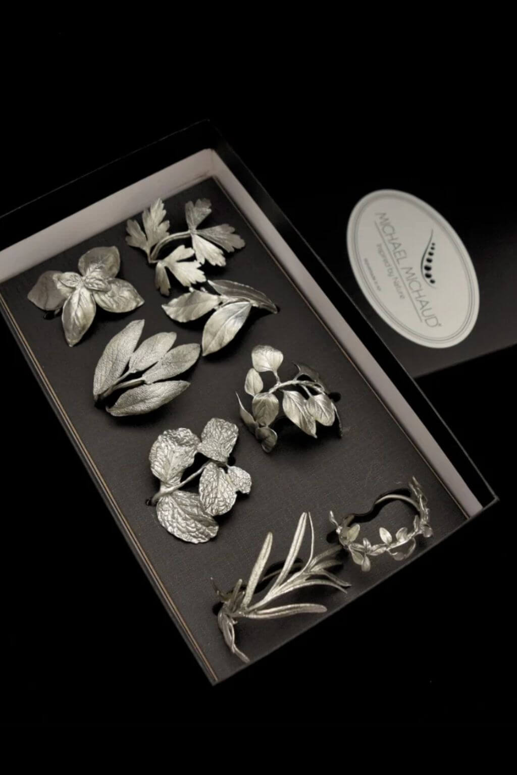 A set of six silver-toned metal leaf napkin rings displayed inside a black presentation box, with a white oval brand tag reading Michael Michaud on a black background.