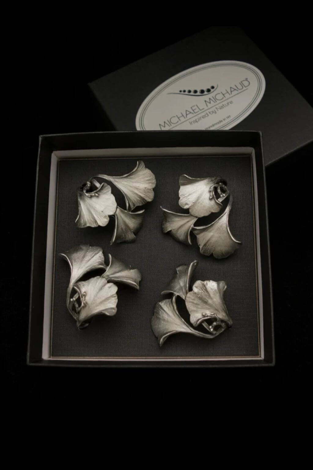 Four pewter inkgo leaf napkin rings displayed inside an open black presentation box, with the branded lid visible behind on a black background.