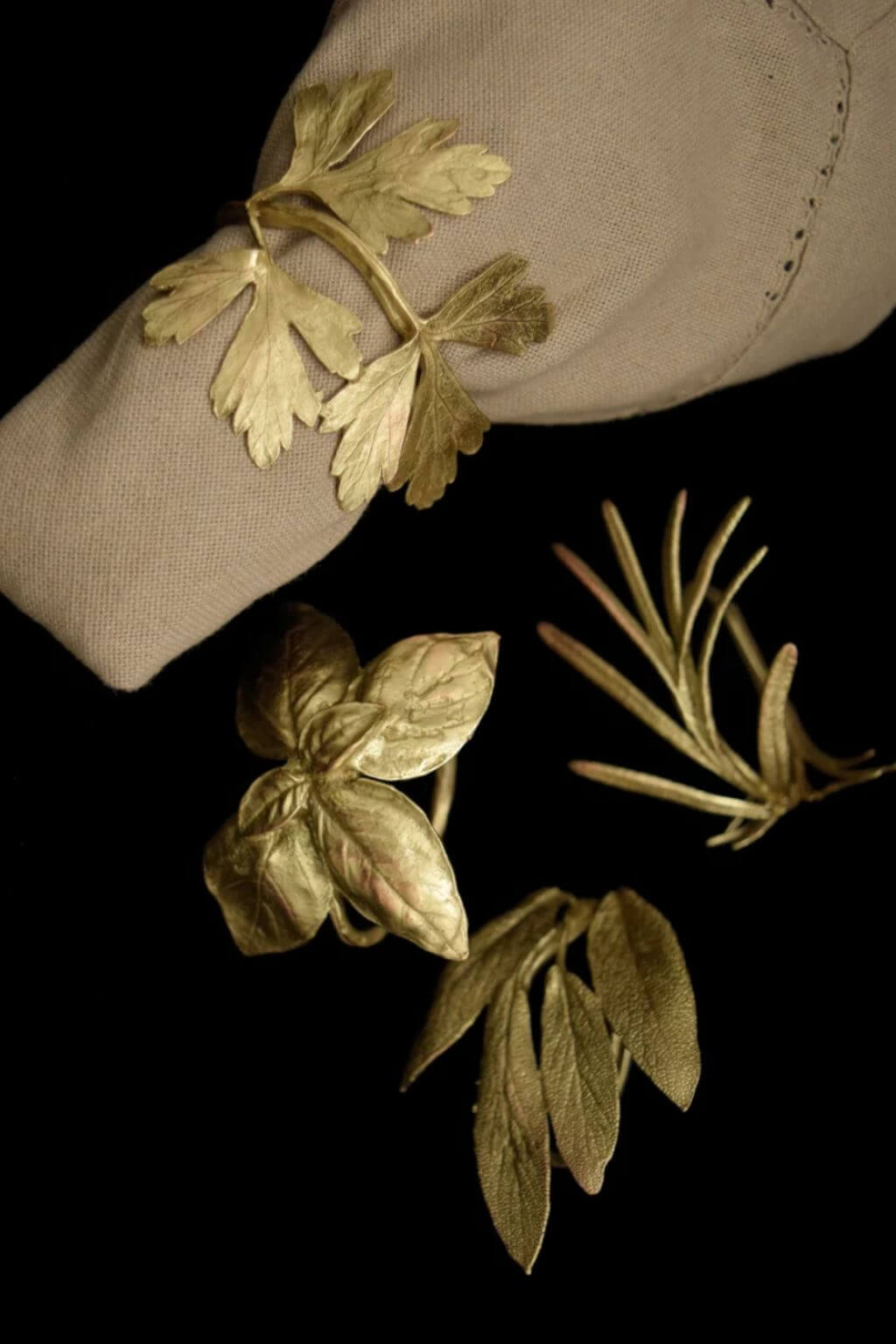 Four gold-toned botanical garden herb napkin rings, displayed against a black background with a beige sleeve in the upper left corner.