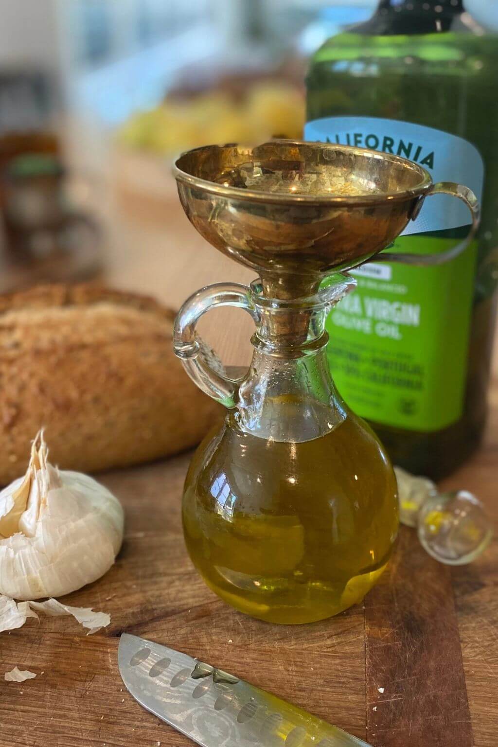 Glass bottle of olive oil with a brass pouring spout on a wooden surface, next to garlic and bread.