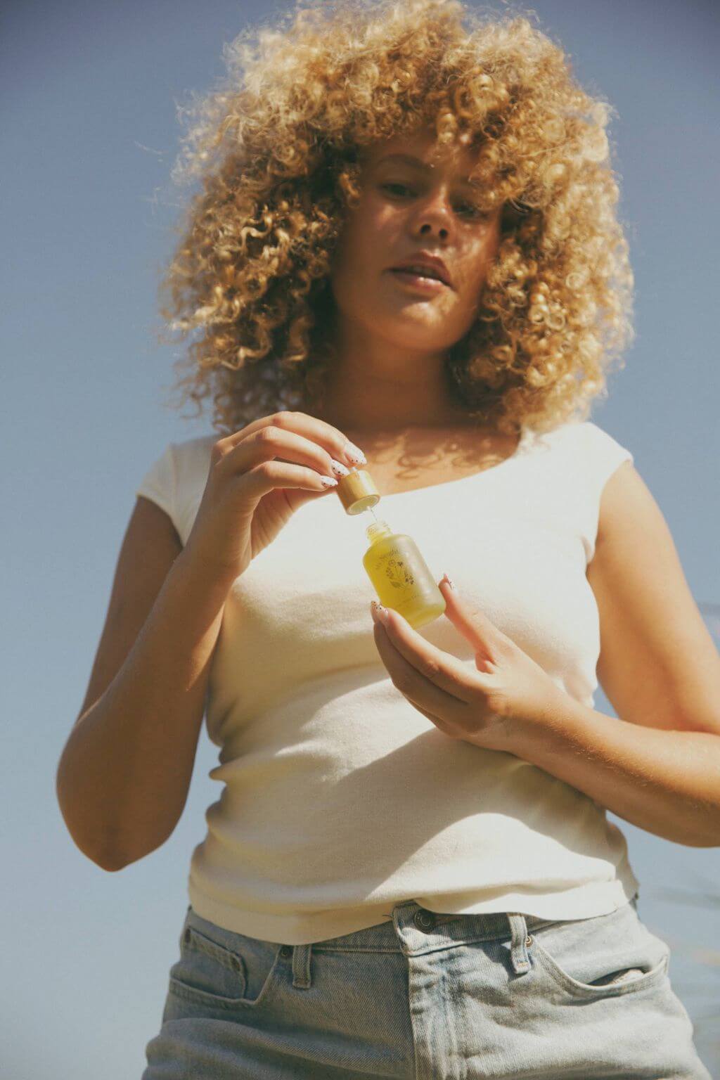 Woman with curly hair holding a dropper bottle of serum against a clear blue sky