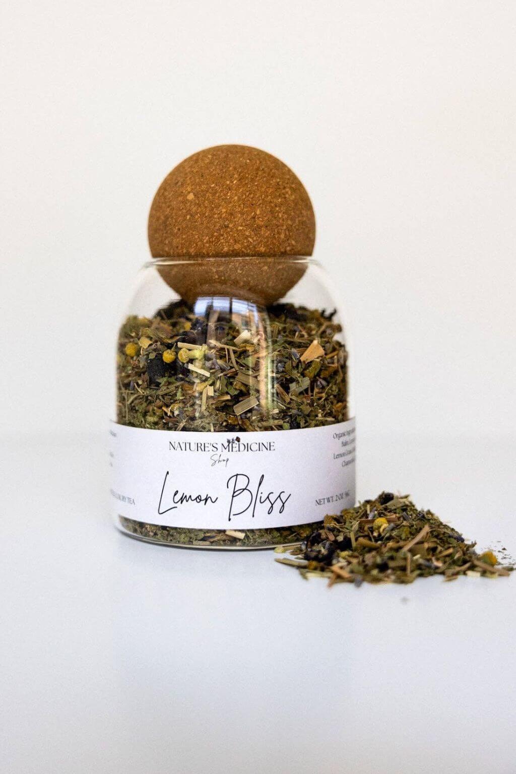 A clear glass jar filled with loose-leaf herbal tea, topped with a round cork stopper and labeled 'Lemon Bliss' by Nature's Medicine, on a clean white background with a small pile of tea beside the jar.
