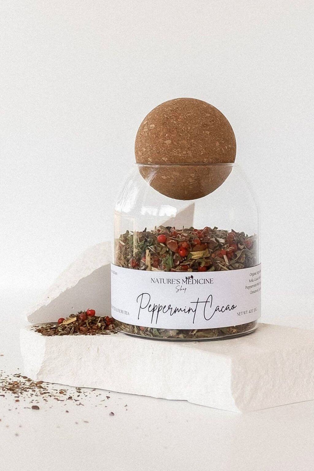 A clear glass jar filled with loose-leaf peppermint cocoa tea, topped with a cork ball lid, displayed on a white stone block against a white background. The label reads 'Nature's Medicine Shop Peppermint Cocoa'.
