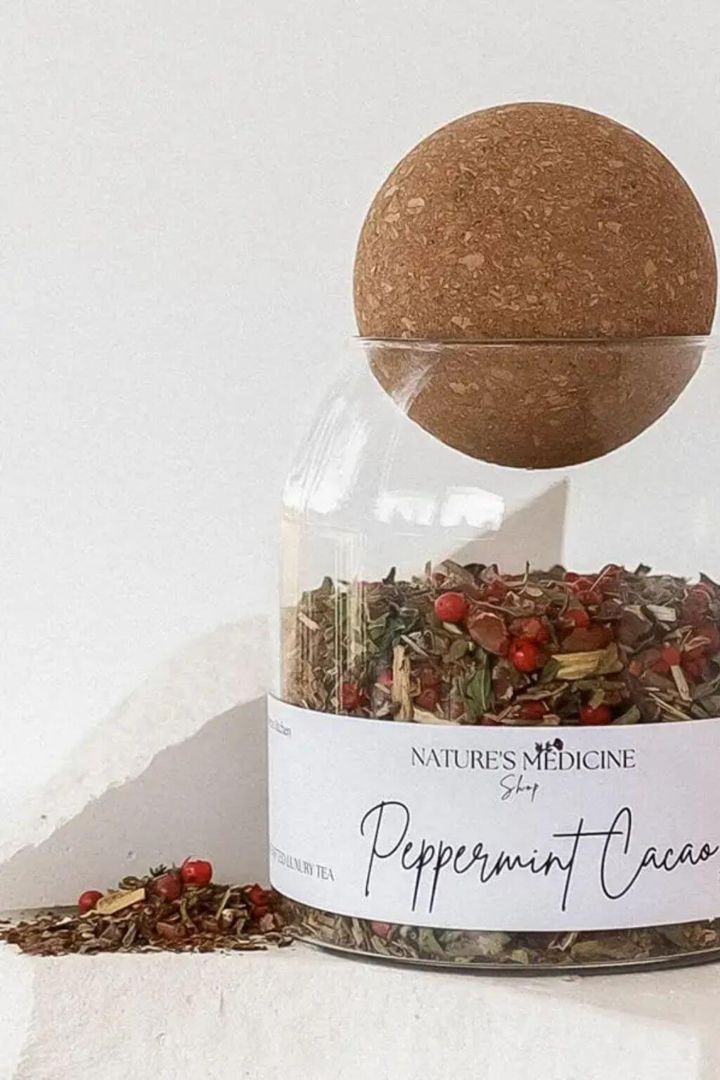 A clear glass jar of Nature's Medicine Shop Peppermint Cacao loose-leaf tea with a cork stopper, shown on a light textured surface with a few loose pieces spilled in front.