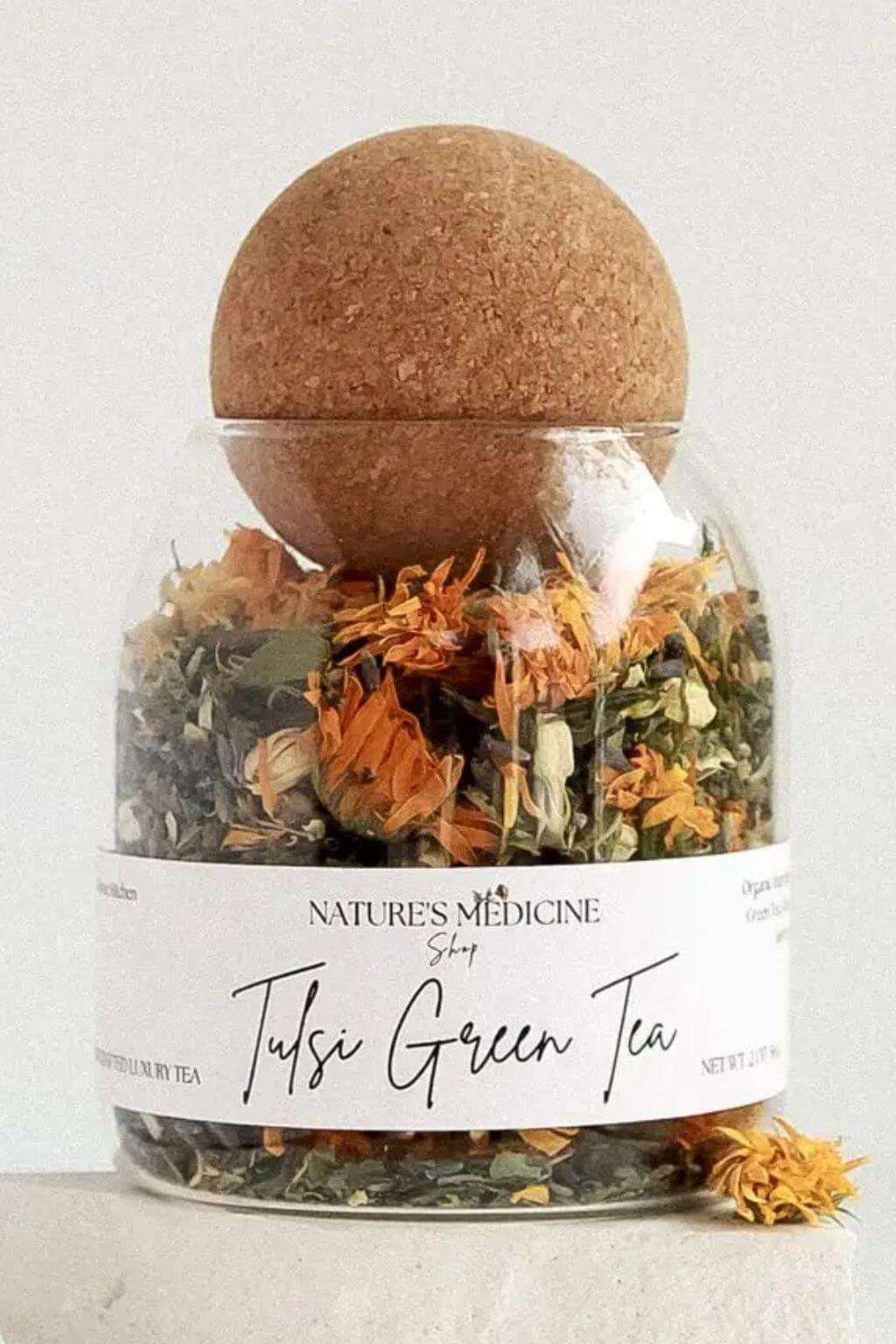 A clear glass jar of Nature's Medicine Shop Tulsi Green Tea filled with dried green and orange botanicals, topped with a brown cork ball, on a light neutral background.