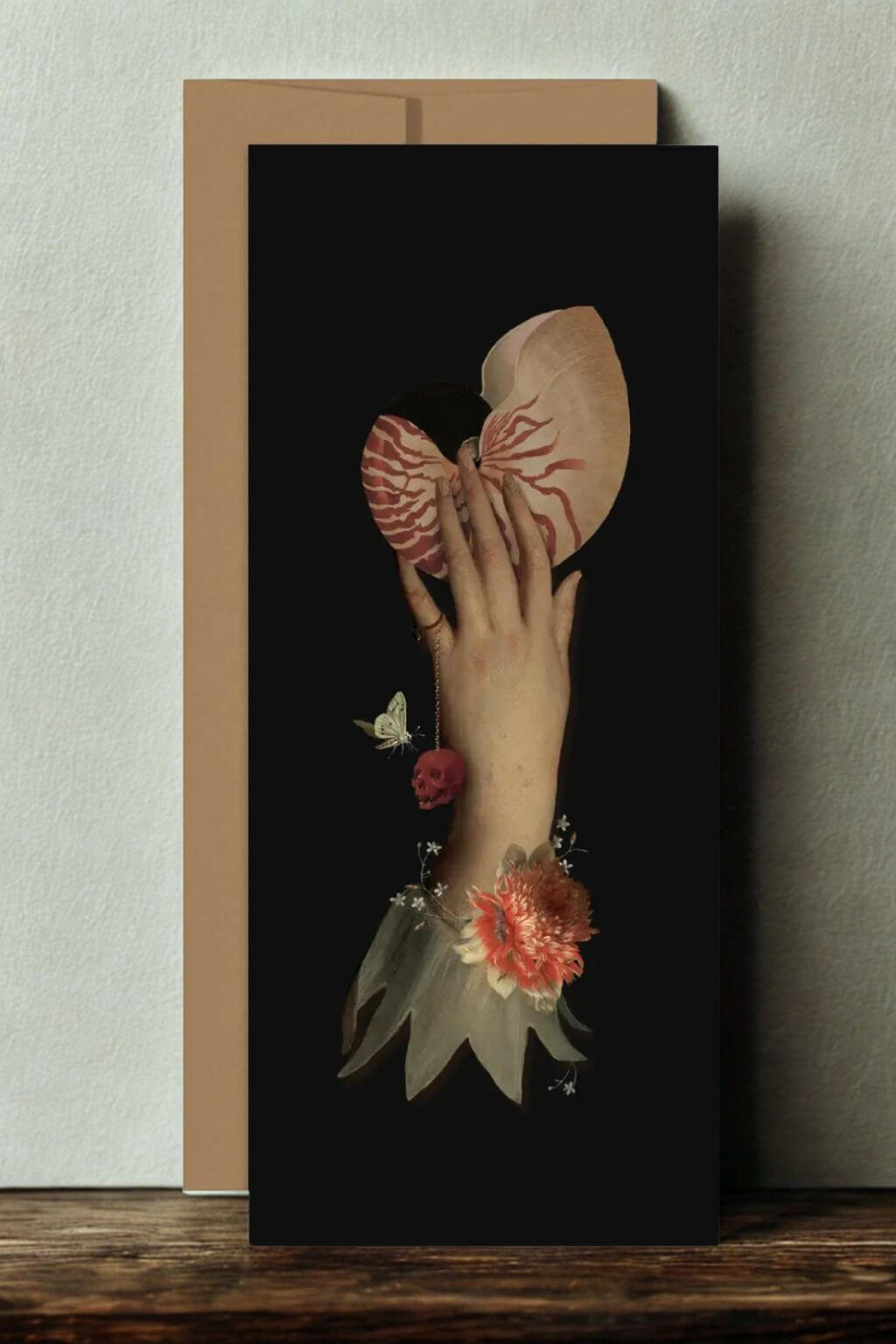 A tall card leaning against a light wall on a wooden surface, with a tan envelope behind it. The card shows a hand holding a nautilus shell with flowers and butterflies around it.