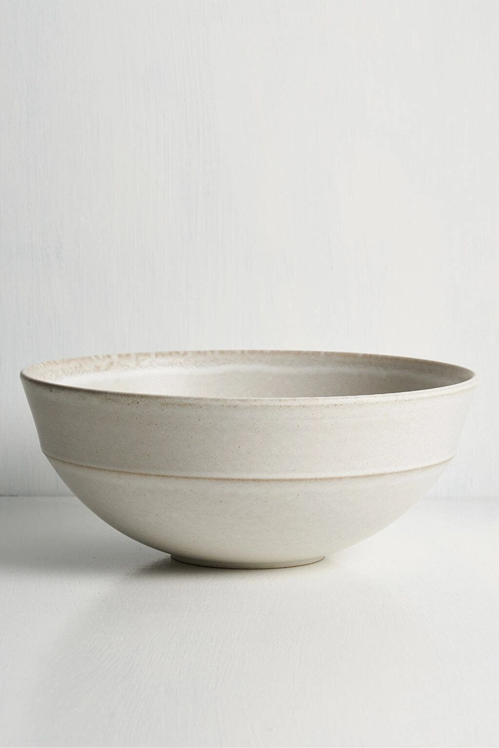 A large, round ceramic bowl with a textured, matte off-white finish, photographed against a clean, light gray background.