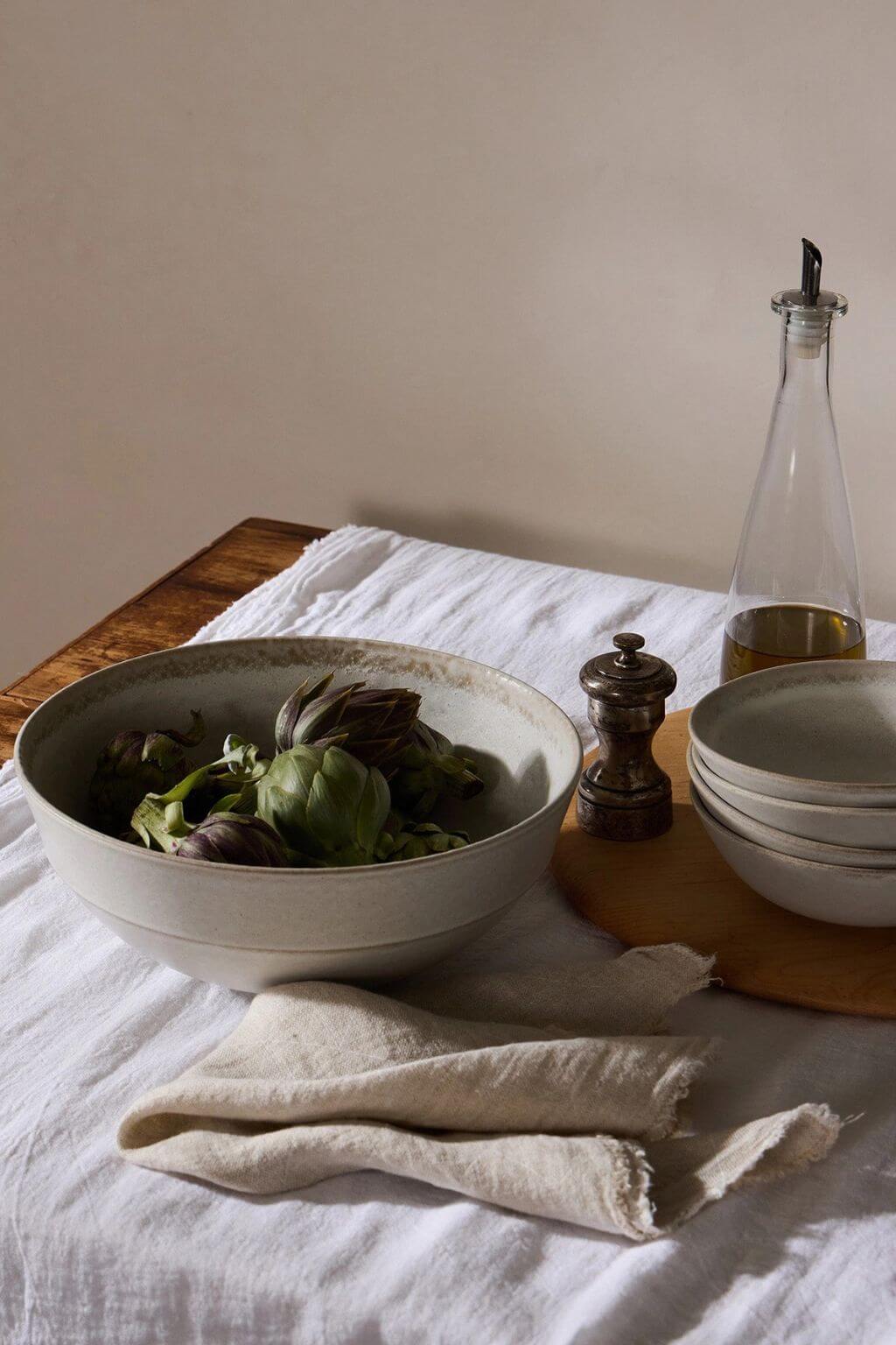 A rustic ceramic bowl filled with green artichokes sits on a white linen tablecloth next to a stack of matching bowls, a wooden pepper mill, and a glass oil bottle.