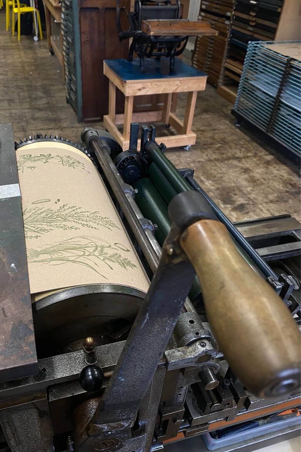 An antique wooden-handled printing press with a sheet of printed paper feeding through rollers, photographed in a workshop or studio interior.