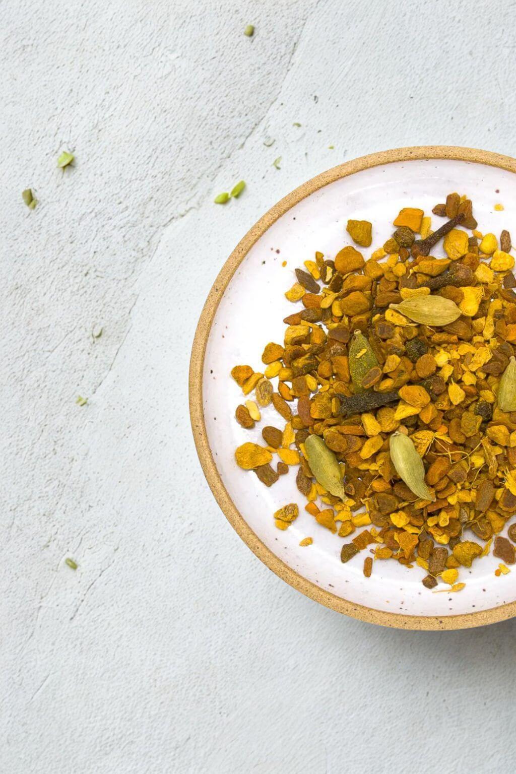 A top-down view of a white ceramic bowl with natural clay rim, filled with a loose herbal tea blend containing yellow turmeric flakes, green cardamom pods, and dried spices, set on a light gray textured surface.