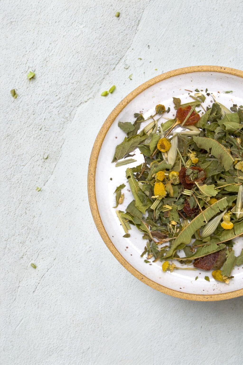 A top-down view of a white ceramic bowl with a natural clay rim, filled with loose-leaf herbal tea containing green leaves, yellow flowers, and small red pieces, set on a textured light gray surface.